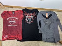 Three women's Harley Davidson shirts laid flat on floor: red short-sleeve, black short-sleeve, grey long-sleeve