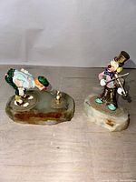 Both clown figurines on individual polished stone bases, one with golf club aiming at a butterfly, the other playing a violin.