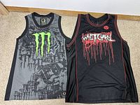 Two sleeveless jersey shirts lying flat, one grey with bright green Monster Energy logo, black mesh pattern, and Fox logo, the other black with red trim and 'White Girl' dripping text with skull icon.