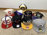 A group photo showing nine sports hats from various teams arranged on carpeted floor against white wall.