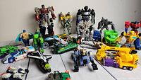 Overview of multiple Transformers toys including robot figures, vehicles, and dinosaur robots arranged on a white wooden surface.