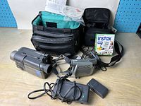 Two camcorders, charging cables, two carrying cases, manual, software CD, and instant film on a wooden surface.