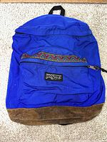 Photo showing the full front view of a vintage blue Jansport backpack with a suede bottom and a colorful geometric trim on the zippered front pocket.