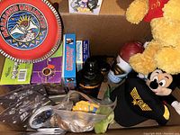 Box containing various plush toys including Winnie the Pooh and Mickey Mouse, a green dragon plush, Darth Vader thermos, and VHS tapes.