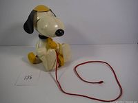 Front angle of the vintage Snoopy pull toy showing Snoopy seated, yellow Woodstock figure on lap, and red pull string laying in front.