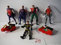 Seven Robin action figures displayed standing and sitting on a white surface, including two figures on red vehicles with 'R' emblem.