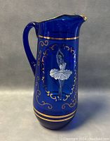 Front side showing standing ballerina with arms raised and detailed gold scrollwork on cobalt blue glass.