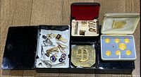 Photo showing various small items including tie pins, a large brass D plate, a black metal box, and a set of gold plated City of London blazer buttons in velvet box.