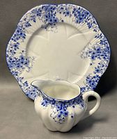 Front view of the plate and creamer showing the blue floral pattern and scalloped edges of the plate.