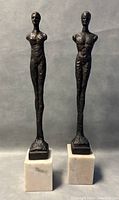 Front view of the two cast metal nude sculptures on marble bases showing full height and form.