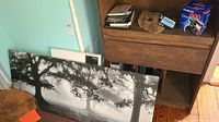 Wide view of the black and white stretched canvas art leaning against wall with wooden furniture and assorted items on top.