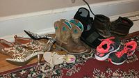 Lot of women's footwear arranged on floor including sandals, heels, western boots, black boots, and running shoes.