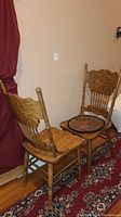 Two vintage wooden chairs with detail carved backs, one with woven seat cushion, on hardwood floor and red rug near red curtain.