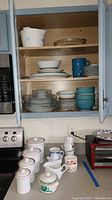 Kitchen cabinet shelves filled with various plates, bowls, mugs, and Corningware containers. Countertop has more ceramic containers with lids, pitchers, and several mugs.