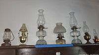 Seven assorted vintage hurricane oil lamps displayed on a shelf showing different styles and sizes.