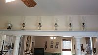 Full view of six vintage hurricane oil lamps lined up on a shelf under ceiling fan. The lamps have a classic glass chimney and base profile with metal burner parts.