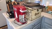 Red Keurig coffee maker and vintage bread box on countertop with waffle iron and griddles behind them.