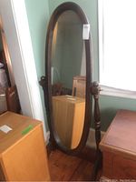 Full view of cheval standing mirror with dark wooden oval frame and base, placed on wood flooring.