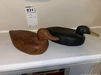 Pair of carved wood ducks on a white surface, natural wood finish duck with red eyes in front and black painted duck behind.
