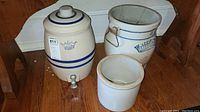 Photo showing all three vintage crocks on wooden floor: tall water cooler crock with spigot and blue crown and stripes, white crock with blue banding, and plain white crock.