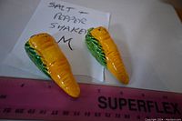 Two yellow carrot ceramic salt and pepper shakers on white paper with handwritten note, next to pink ruler for scale.