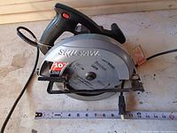 Top and side view of the Skilsaw circular saw showing the blade and power cord along with a tape measure for scale.
