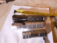 Top view of three flashlights laid next to a measuring tape showing length approximately 6 inches for the Husky flashlight and smaller sizes for the silver flashlights.
