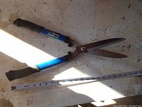 Blue Fiskars Garden-Rite hedge shears laid open on surface, showing blades and rubber grips on handles alongside measuring tape.