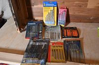 Assorted jig saw blades and masonry drill bits displayed on a table showing various packaging types including plastic, cardboard, and blister packs.