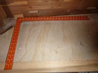 Full view of the Mayes rafter square, showing the overall shape and red-orange metal with measuring markings.