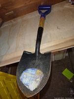 Photo showing full shovel with yellow and black handle, blue D-shaped grip, and metal blade with a sticker residue.