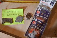 Overall view of three spark plug gap tools and six unopened packages of NGK Iridium spark plugs on a wooden surface with a measuring tape in background