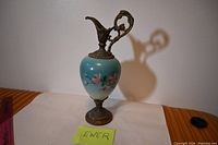 Full frontal image showing the Victorian style ewer with blue body, floral painted design, ornate bronze/brass base and handle, standing upright on a white surface.