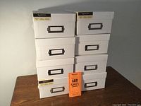 Eight white rectangular memory boxes stacked in two columns on a wooden table, showing front metal label holders and brand labels.