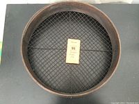 Top view photo showing the circular bentwood frame and wire mesh screen with a label attached in the center.