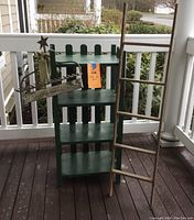Green painted vintage bookcase with four shelves and a green picket fence back design, and a tall wooden ladder leaning beside it, along with a rustic holiday sign on the left side.