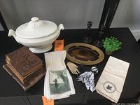 Lot arranged on black surface; large white ironstone tureen with lid, two wooden candle holders, stack of six antique books, two cloth tea towels, brass tray, small vintage glove and paper cut-out figure.