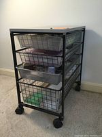 Full view front angle of black Elfa storage cart with four wire mesh drawers containing office supplies and a gray top.