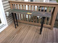 Full view of folk art table on wooden porch showing narrow table made with a wooden shutter top and black legs.