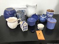 Full lot showing assortment of blue and white ceramics including pitchers, crocks, Delft house, lidded jars, plates, and decorative items arranged on black surface.