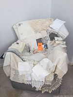 Image showing assorted vintage linens draped over a chair, including multiple pieces of crocheted lace, embroidered table linens, and white napkins.
