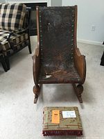 Front view of the oak rocking chair showing the carved wood frame and worn leather sling seat and back, with a small footstool in front.