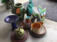 Wide view showing blue bird bath, green strawberry pot with multiple pockets, several terracotta and ceramic flower pots stacked in plant trays, and bags of potting soil on concrete floor.