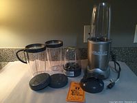 Full view of blender base with attached blending cup, two additional blending cups, small processing cup with blade, gray lids, instruction manual and recipe book