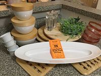 Full view of various kitchen items including stacked white bowls with wooden lids, small white dipping bowls, a long white serving dish, salt and pepper shakers, decorative greenery in container, three wooden lazy Susans stacked, and two wooden trivets beneath dishes.