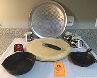 Overview of pizza stone, perforated pizza pan, two cast iron skillets, pizza cutter, three mugs and hotpad handle cover arranged on table.