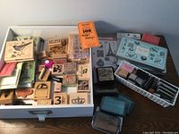 Top-down image showing a white tray containing many wooden-block mounted rubber stamps, including bird, crown, and number designs. Several ink pads and clear stamps are to the side.