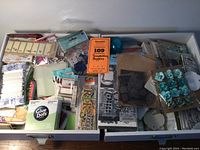 Overview photo showing assembled scrapbooking and assemblage art supplies in two cardboard trays or boxes, displaying lace trims, flowers, adhesives, labels, and other embellishments.