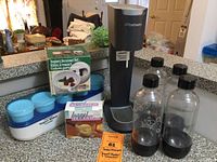 SodaStream machine, four bottles including three black cap and one blue cap, yogurt strainer set with blue lid containers, and a box of bagel holders on kitchen counter.