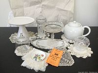 Wide shot showing entire tea party set including ceramic items, glass dishes, and lace doilies arranged on table.